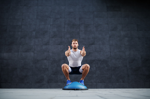 Front view of handsome caucasian muscular bearded man doing squat exercise on bosu ball. In background is gray wall.