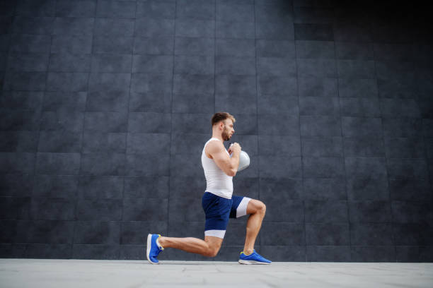 homme caucasien musculaire fort beau dans des shorts et des t-shirt faisant des fentes et retenant la cloche de bouilloire. en arrière-plan est mur gris. - core workout photos et images de collection