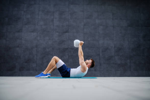 Side view of handsome caucasian muscular man lying down on mat and lifting kettle bell. Side view of handsome caucasian muscular man lying down on mat and lifting kettle bell. apple core stock pictures, royalty-free photos & images