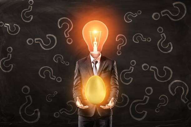 istock Man in suit with lightbulb instead head, levitating big shiny golden egg above palms, standing against black wall with pattern of question marks on it. 1179560796
