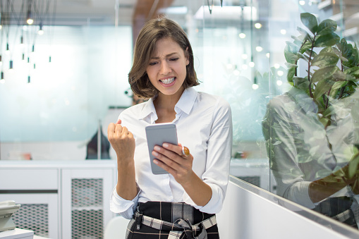 Excited young business woman receiving good news on smartphone in an office
