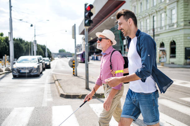 Young man and blind senior with white cane walking in city, crossing street. A young man and blind senior with white cane walking in city, crossing street. blindness stock pictures, royalty-free photos & images