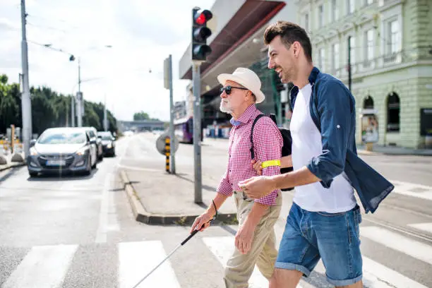 A young man and blind senior with white cane walking in city, crossing street.