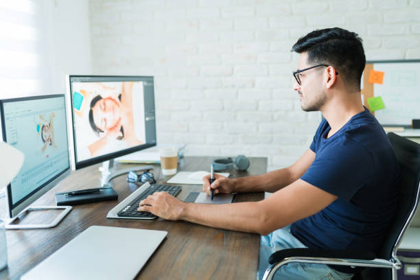 Handsome Young Male Editor Using Graphic Tablet At Desk Side view of attractive Hispanic retoucher editing photo while working from home editor photos stock pictures, royalty-free photos & images