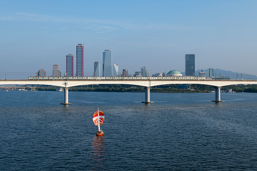 View from the Yanghwa Bridge of a sailboat sailing on the Han River in Seoul, South Korea. A train is crossing a railway bridge.