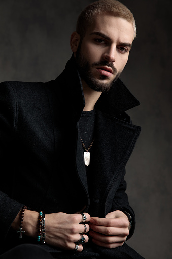 attractive young model wearing black longcoat, holding hands and arranging coat, sitting on grey background in studio