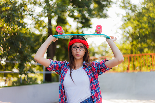 Funny young hipster woman dressed in stylish clothes holds skateboard in her hand in skatepark at bright sunny day. Summer lifestyle image of trendy pretty young girl