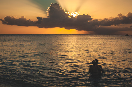 A young couple holding each other while bathing in the tropical sea watching the sun low on the horizon. It could be either sunrise or sunset. The ocean is calm.