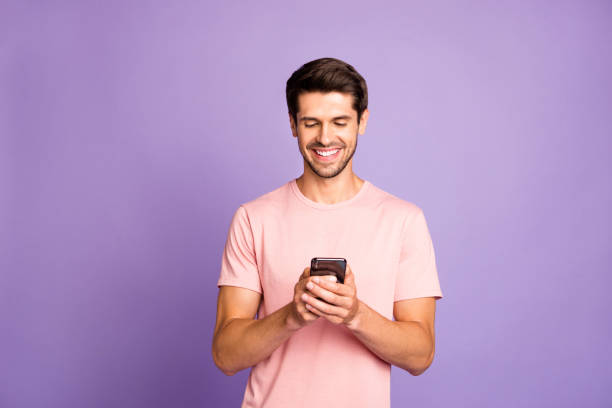 Portrait of his he nice attractive cheerful cheery guy wearing pink tshirt holding in hands using 5g app cell modern technology isolated over violet purple lilac pastel color background Portrait of his he nice attractive cheerful cheery guy wearing pink tshirt holding in hands, using 5g app cell modern technology isolated over violet purple lilac pastel color background email campaign photos stock pictures, royalty-free photos & images