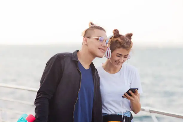 Photo of Youth summer time. Stylish hipster couple in love strolling along the beach promenade and listen to music with headphones.