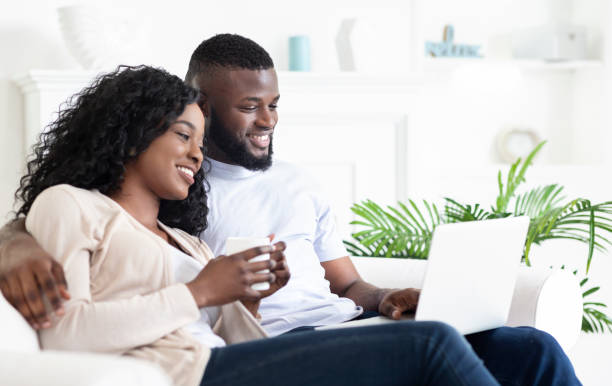 istock Cheerful black couple sitting on couch looking at laptop screen 1179597578