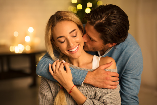 Tender moment. Man kissing wife in front of Christmas tree in cozy living room