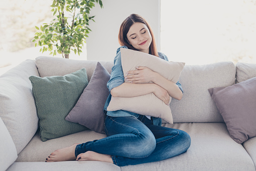 Photo of pretty lady holding big pillow close to chest closing eyes glad get rest, after hard working week sitting sofa wearing jeans clothes apartment indoors
