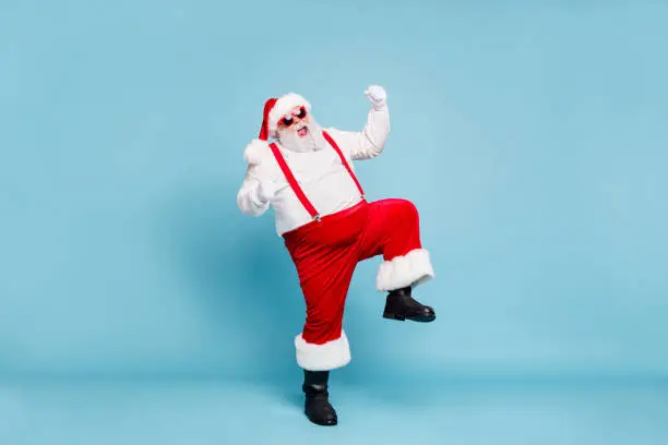 Photo of Full body photo of crazy funny funky fat santa with big belly raise fists scream yeah enjoy christmas party wear red pants style stylish boots overalls spectacles isolated over blue color background