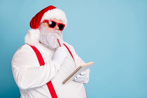 Close up photo of focused funny funky fat santa claus hold notebook, think about wish list plan decide what choose look thoughtful wear stylish trendy suspenders isolated over blue color background