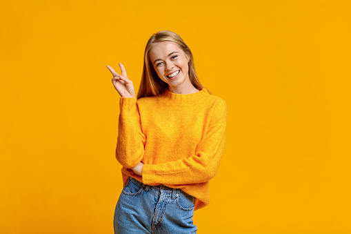 Smiling redhead teenager showing peace gesture and posing to camera, orange studio background with free space