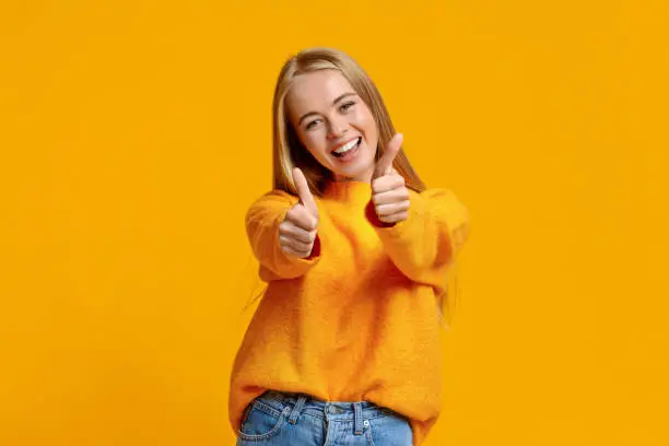 I like it. Portrait of joyful teenage girl showing thumbs up and smiling at camera, orange background with copy space