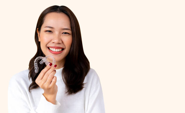 close up (isolated) young beautiful asian woman smiling with hand holding dental aligner retainer (invisible) on cream background of dental clinic for beautiful teeth treatment course concept close up (isolated) young beautiful asian woman smiling with hand holding dental aligner retainer (invisible) on cream background of dental clinic for beautiful teeth treatment course concept dental aligner photos stock pictures, royalty-free photos & images