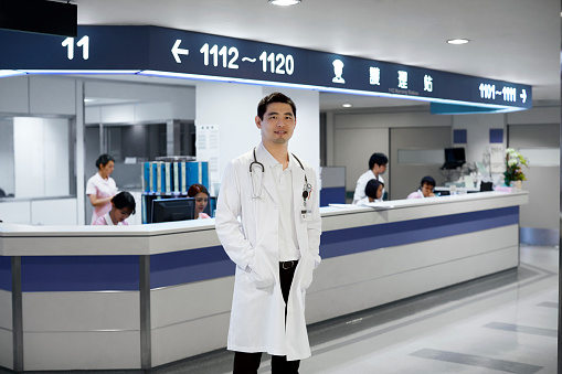 Portrait of male doctor with hands in pockets at hospital. Confident healthcare worker standing against colleagues working at reception. Medical professional is wearing lab coat.
