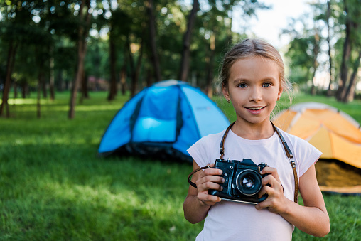smiling child holding digital camera near camps in park