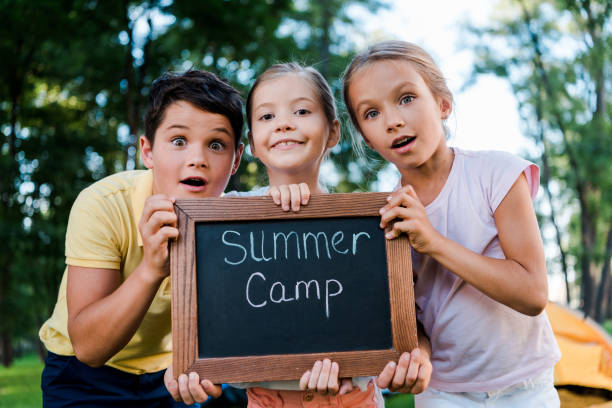 niños sorprendidos sosteniendo tablero de tiza con cartas de campamento de verano - campamento de verano fotografías e imágenes de stock