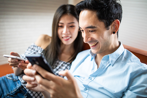Young Asian couple relaxing at home, sitting on sofa and looking at smart phone