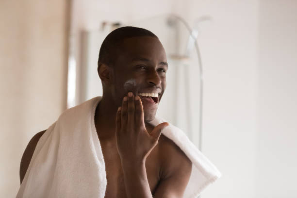 szczęśliwy uśmiechnięty afrykański mężczyzna pochodzenia etnicznego stosujący balsam nawilżający na policzek. - men shaving human face bathroom zdjęcia i obrazy z banku zdjęć