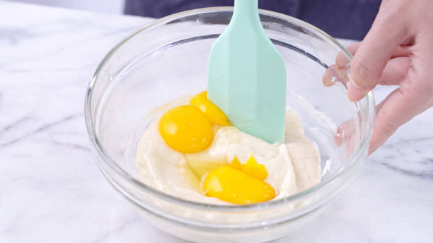 Mixing egg yolk into cake batter with green rubber spatula mixer tool stirring until smooth and blend well in a glass bowl.