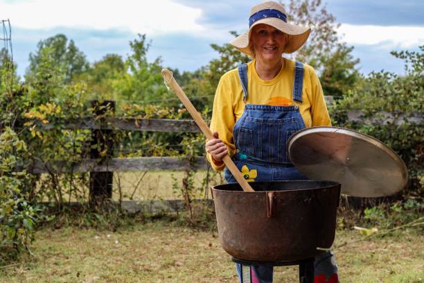 istock Middle aged woman cooking over an open fire in a large black cast iron kettle, holding a large wooden paddle 1179626065