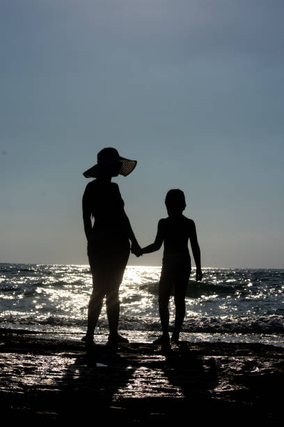 Silhouette of a woman wearing a sunhat and holding the hand of a young boy at the beach