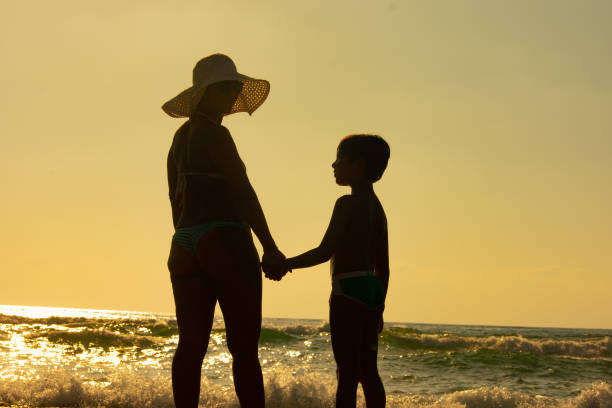 Silhouette of a woman wearing a sunhat and holding the hand of a young boy at the beach