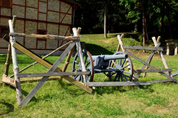 istock Single massive grey cannon replica on two rusty wheels with base and barrel  visible stanting next to an ad hoc wooden barricade in the middle of a lush field, pastureland, or meadow seen in Poland 1179646380