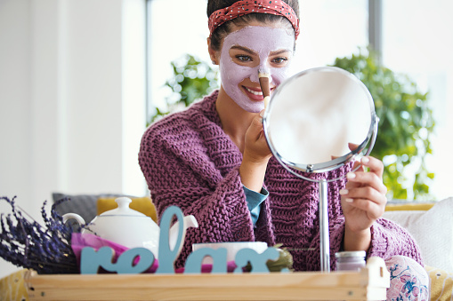 Young woman applying facial mask. She's sitting on her bed looking at mirror and taking care of here skin during weekend morning.