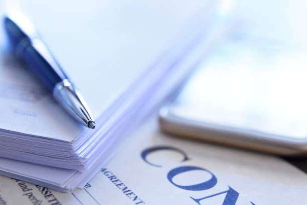 A ballpoint pen rests on top of a stack of documents and an unsigned contract as a mobile phone sits in the background. Photographed with a very shallow depth of field.