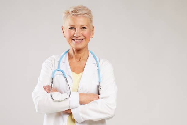 portrait of positive mature female general practitioner with stethoscope standing against white background and crossing arms on chest - healthcare and medicine nurse doctor general practitioner imagens e fotografias de stock