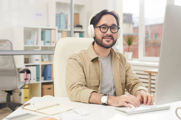 istock Content handsome young Asian designer in headphones sitting at table and typing on keyboard while enjoying music during work 1179662199