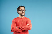 Cheerful dreamy young Asian man with beard and mustache wearing stylish sweater standing against blue background and looking away while being dreamy