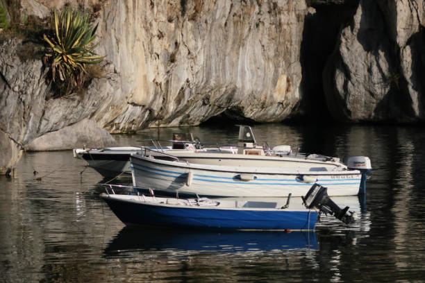 Maratea, Italy - September 6, 2018:  Three small motorboats appear in succession moored to anchors inside a cavernous stone grotto lagoon marina under soft natural lighting in coastal Maratea, Italy.  The three boats appear close-up at lower center of the horizontal frame, all pointed toward the left, one in front of the other.  The water is lightly rippled, displaying abstract reflections of the boats and surrounding stone walls of the grotto.  Softly focused, with an atmospheric feel to the image.