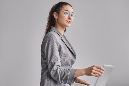 Side view of young woman in glasses and elegant jacket holding laptop and looking away while working in office against gray background