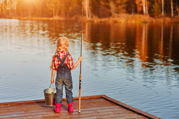 niña con una caña de pescar y un cubo vino a pescar y mira al estanque. vista posterior - peto fotografías e imágenes de stock