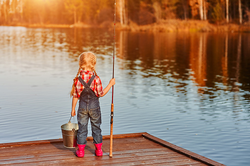 little girl with a fishing rod and a bucket came fishing and looks at the pond. Back view