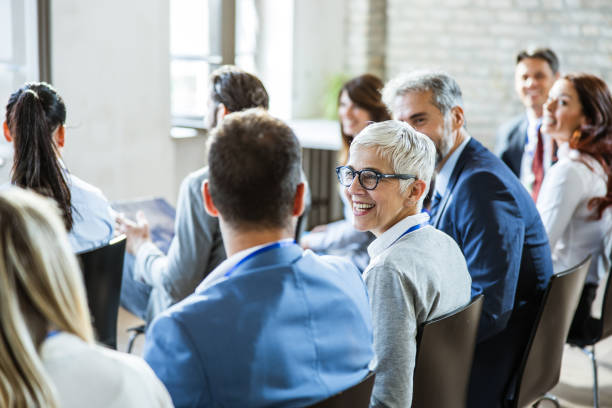 Happy senior businesswoman talking to her colleagues on seminar in the office. Large group of entrepreneurs on a seminar in board room. Focus is on happy mature woman communicating with her colleagues. blue business stock pictures, royalty-free photos & images