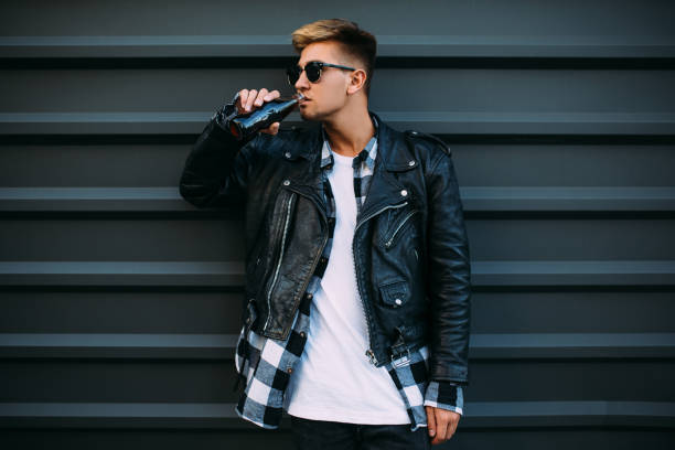 istock A handsome man in sunglasses, in a leather black jacket posing and drinking a drink near a black wall 1179734429