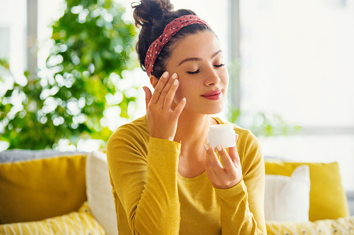 Young woman with eyes closed applying face cream.