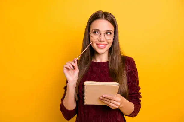Portrait of interested lady holding pen and notebook writing check plan to do list with eyewear eyeglasses wearing burgundy sweater, isolated over yellow background