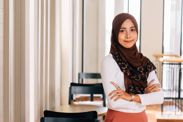 unga lyckliga och framgångsrika sydostasiatiska islamiska affärskvinna med vapen korsade i business corporate building inställning tittar på kamera - business malaysia bildbanksfoton och bilder
