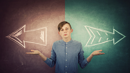 Perplexed teenage boy spread outstretched arms, balancing hands gesture, two options to choose right or left, decision concept. Split blackboard and arrows going in two different ways.