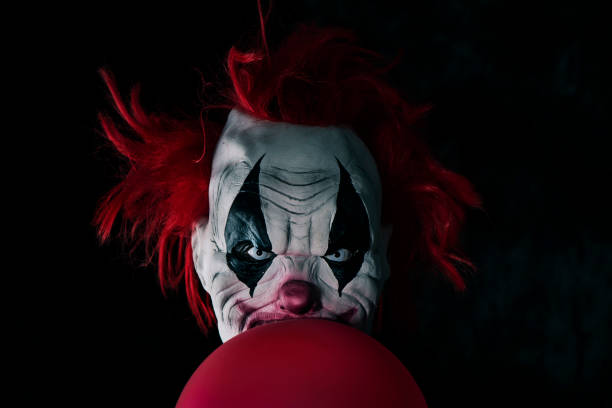 scary evil clown with a balloon closeup of a scary evil clown with red hair and white eyes, staring at the observer, with a balloon in front of him, against a black background face paint halloween adult men stock pictures, royalty-free photos & images