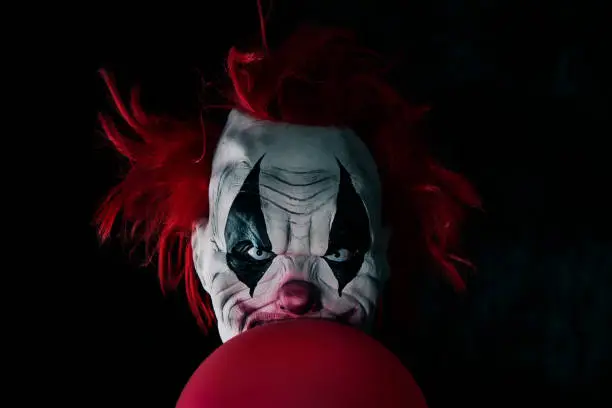 closeup of a scary evil clown with red hair and white eyes, staring at the observer, with a balloon in front of him, against a black background
