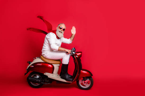 envejecido santa hombre con el pelo gris movimiento fiesta por ciclomotor vintage usar jersey blanco y pantalones de fondo rojo aislado - santa claus waving christmas photography fotografías e imágenes de stock